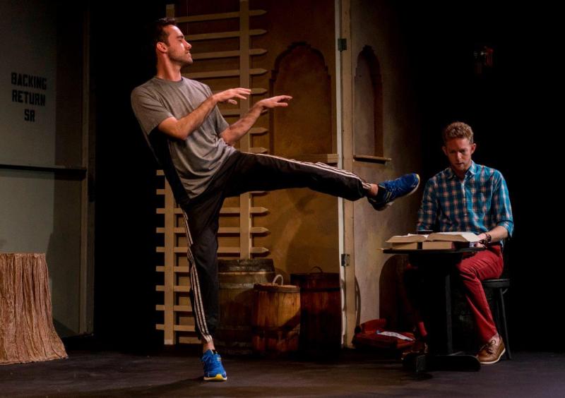 Man in grey shirt and blue pants dances on stage while other man looks on.