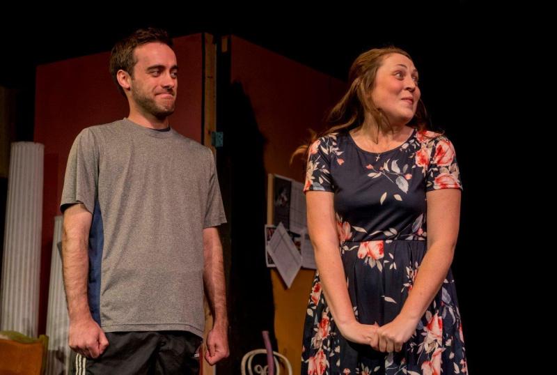 Smiling man in grey shirt stands on stage next to smiling woman in flowered dress.
