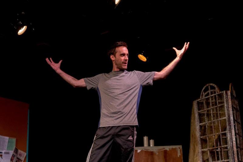 Man in grey shirt with both arms up acting out a scene  on stage.