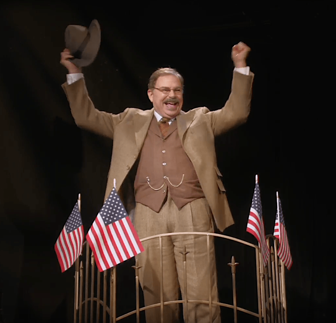 Actor Phil Johnson as President T. Roosevelt smiling on train car surrounded by American flags