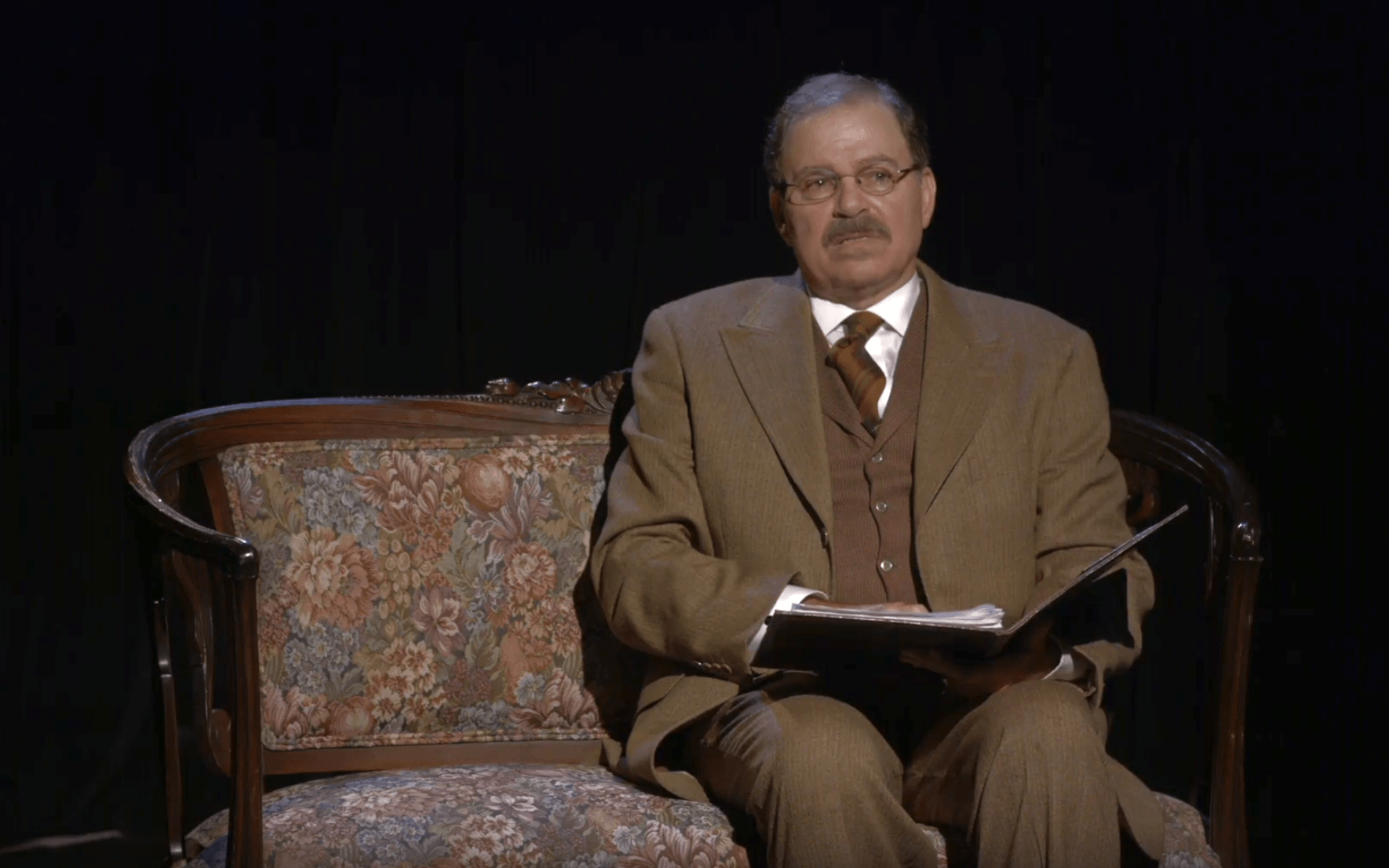 Actor Phil Johnson as President Theodore Roosevelt sitting on a sofa reviewing papers.