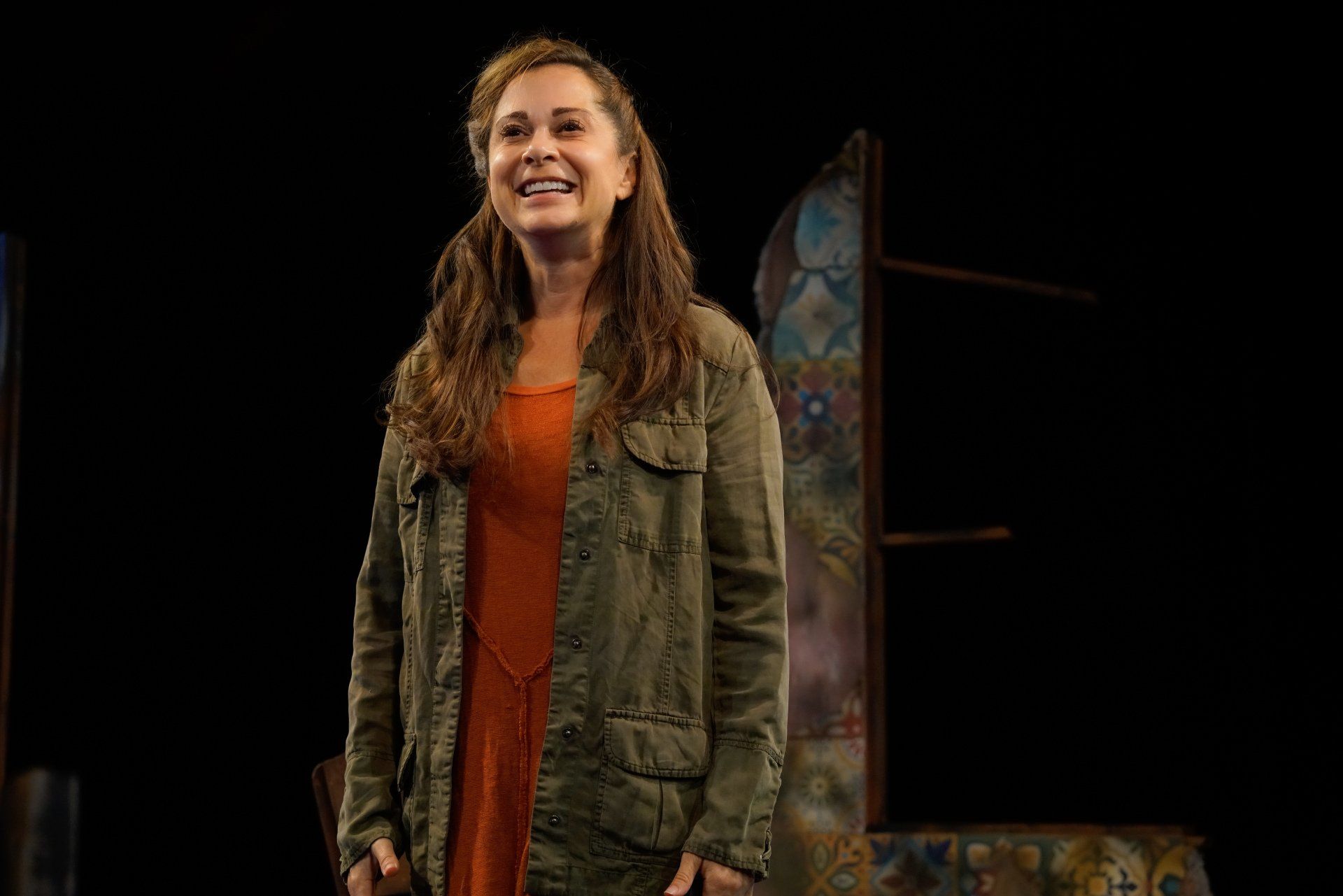 Young Mahshid, smiling, wearing a green jacket and orange sweater.