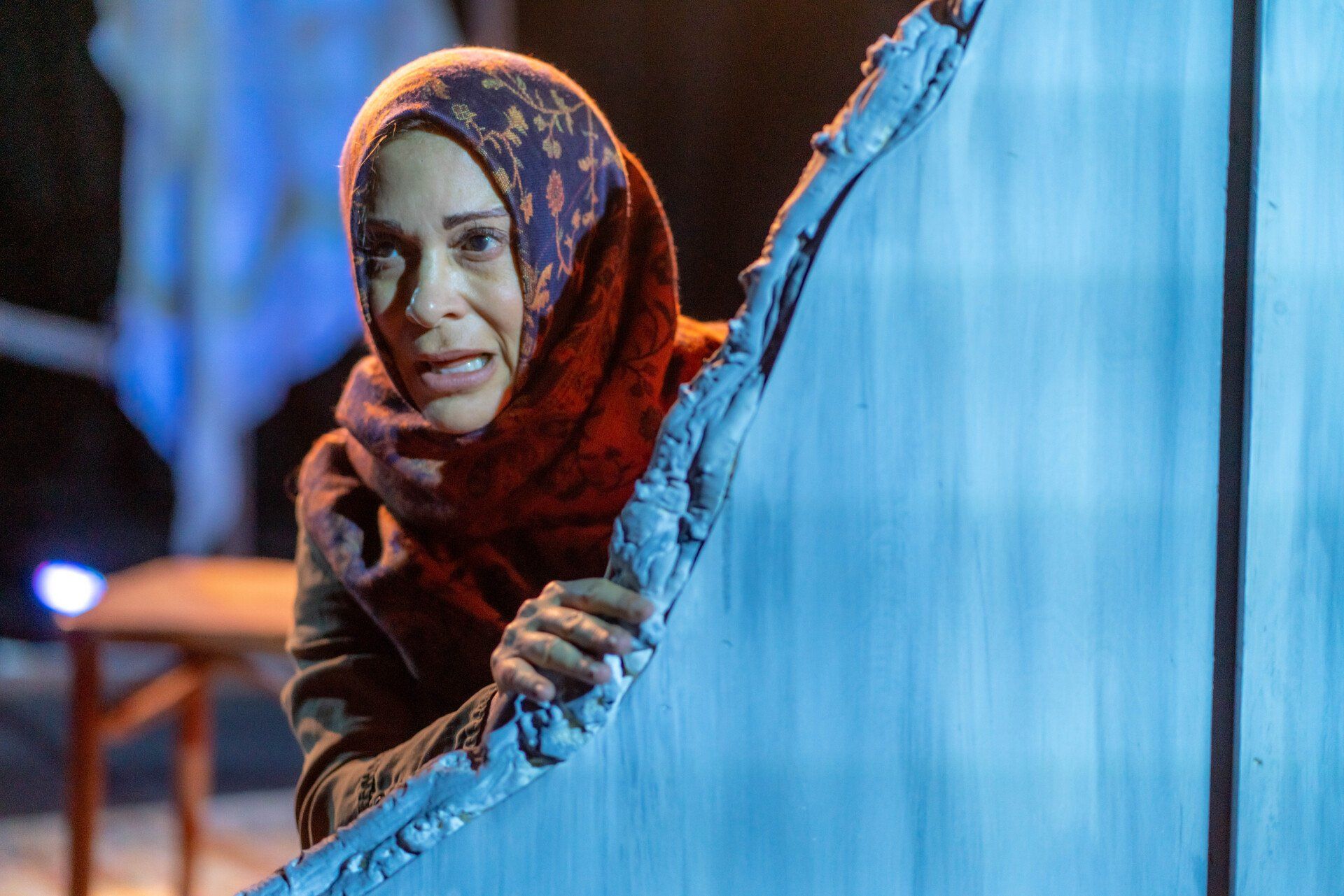 Young Mahshid wearing headscarf and green jacket peering around a wall.