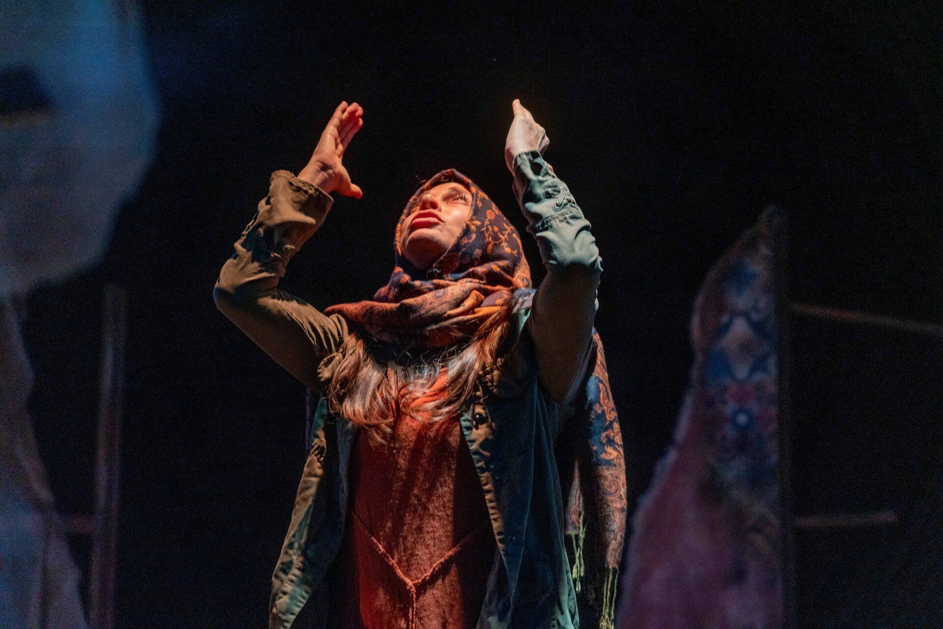 Young Mahshid in orange sweater, green jacket, and headscarf, looking up with hands in the air.