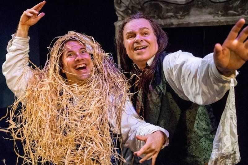 Man wearing a straw headpiece and man in long wig and black vest cavorting onstage.