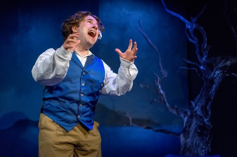 Man in wig, blue vest and ribbon singing in a scene.