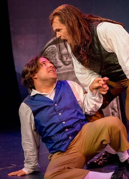 Man in blue wig and vest and man in wig and black vest in tender scene.