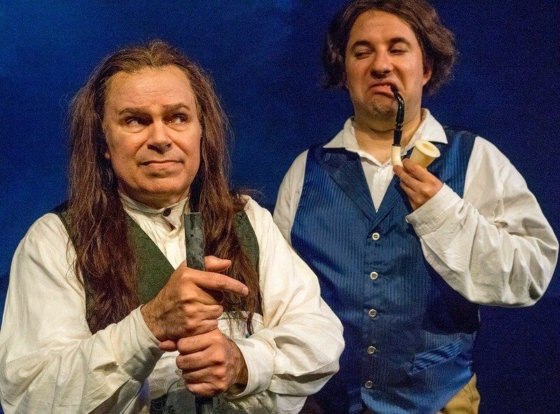 Man in long wig, black vest, and holding broom handle and man in blue vest, wig, and holding a pipe.