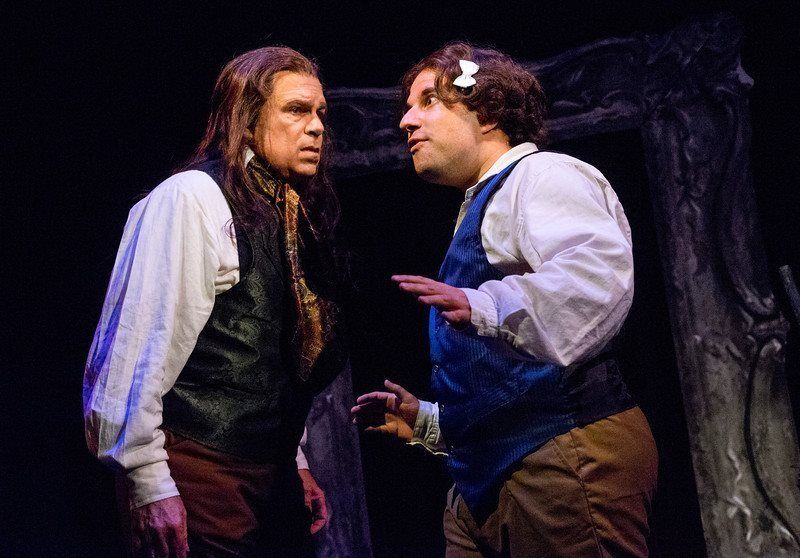 Man in long wig and black vest and man in blue vest, wig, and hair ribbon having a discussion.