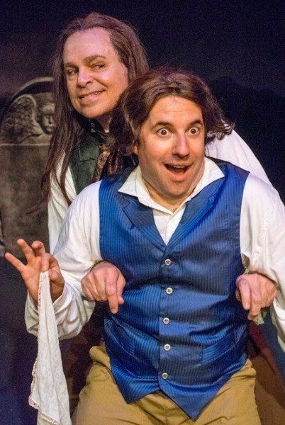 Man in long wig, vest and holding scarf and man in blue vest, wig, and hair ribbon looking gleeful.