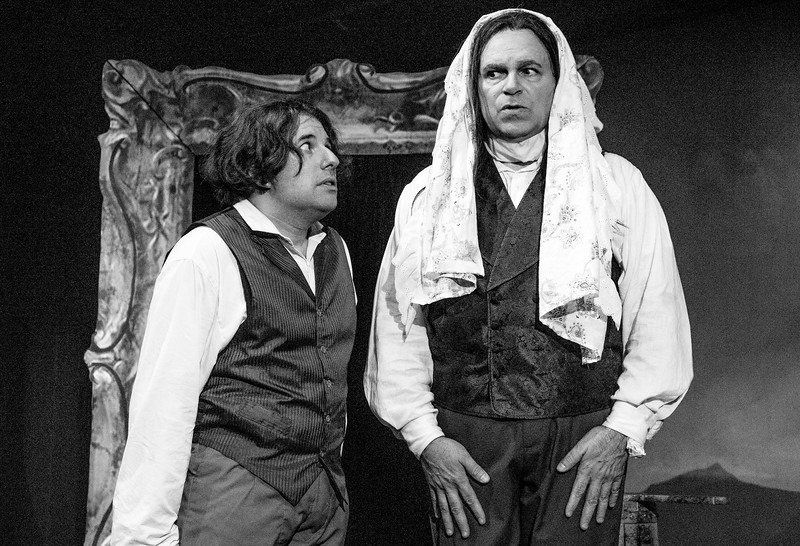 B& W of man in vest and wig and man in wig, vest and wearing a headscarf looking concerned.
