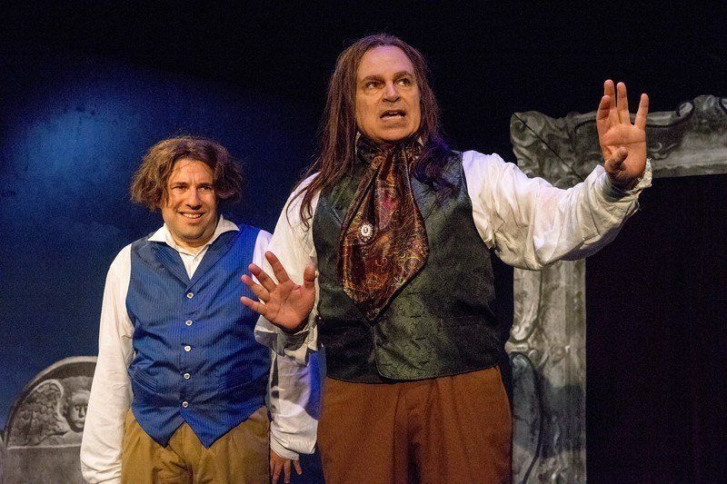 Man in blue vest and wig and man in long wig and black vest sharing a scene.