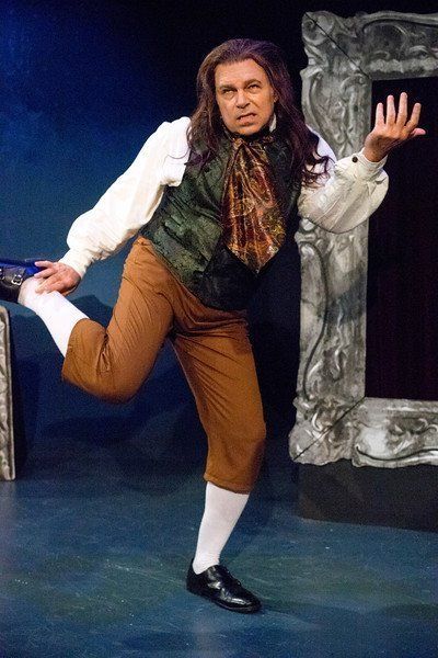 Man in long black wig and black vest dancing in a scene.