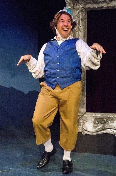 Man in wig, blue vest, and headpiece dancing in a scene.