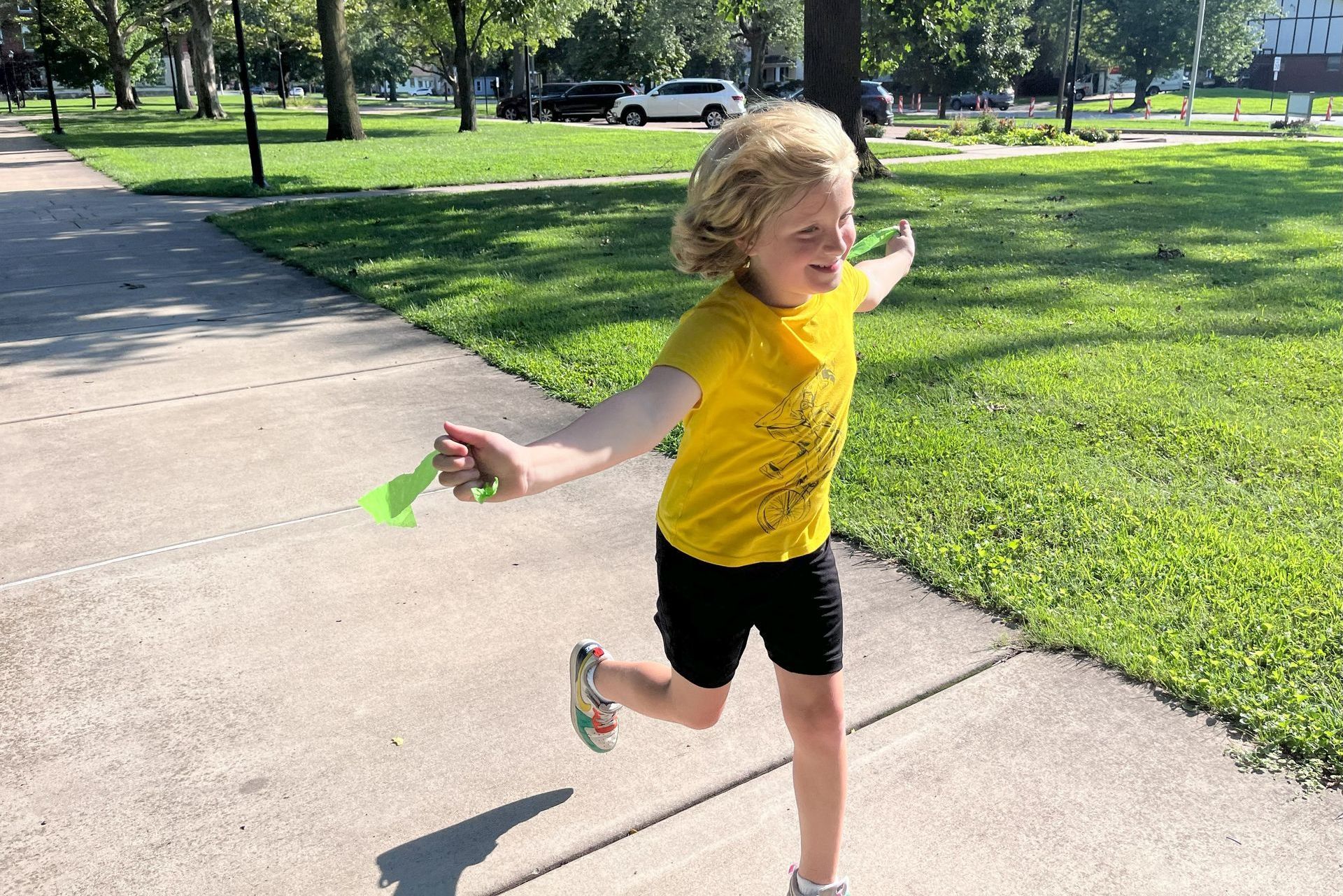 A child with blonde curly hair runs joyfully along a sidewalk in a park, holding a green toy.