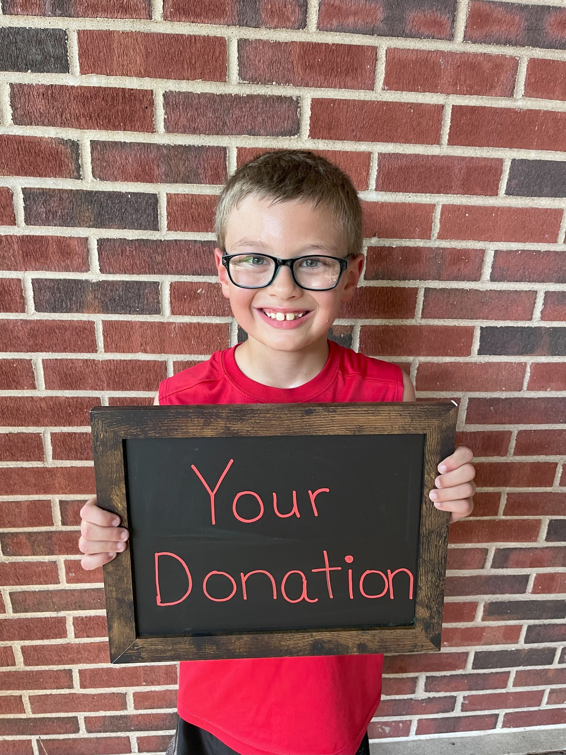 Young boy with glasses smiles holding a chalkboard sign that says 