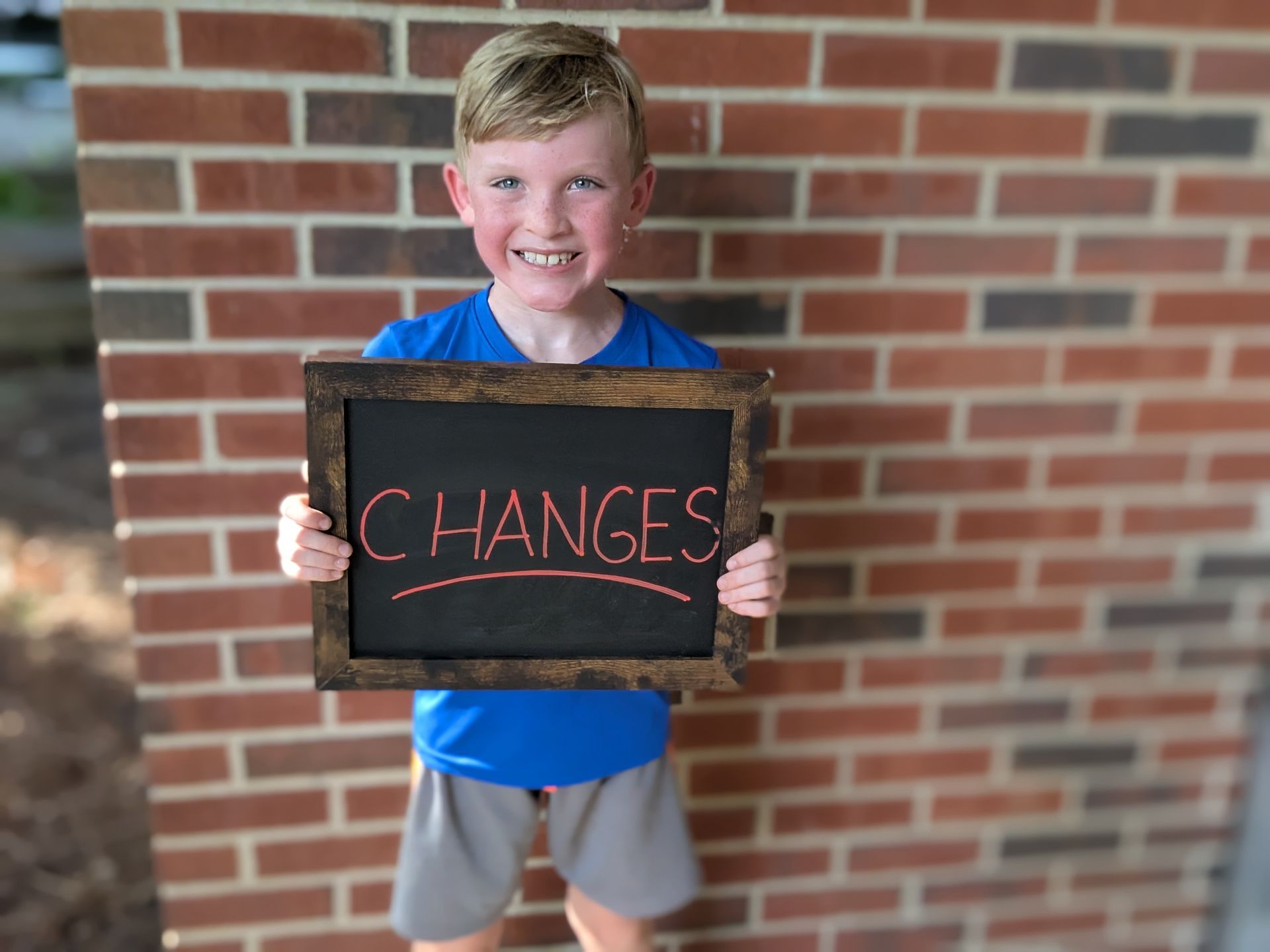 A young boy smiles, holding a chalkboard sign with the word 