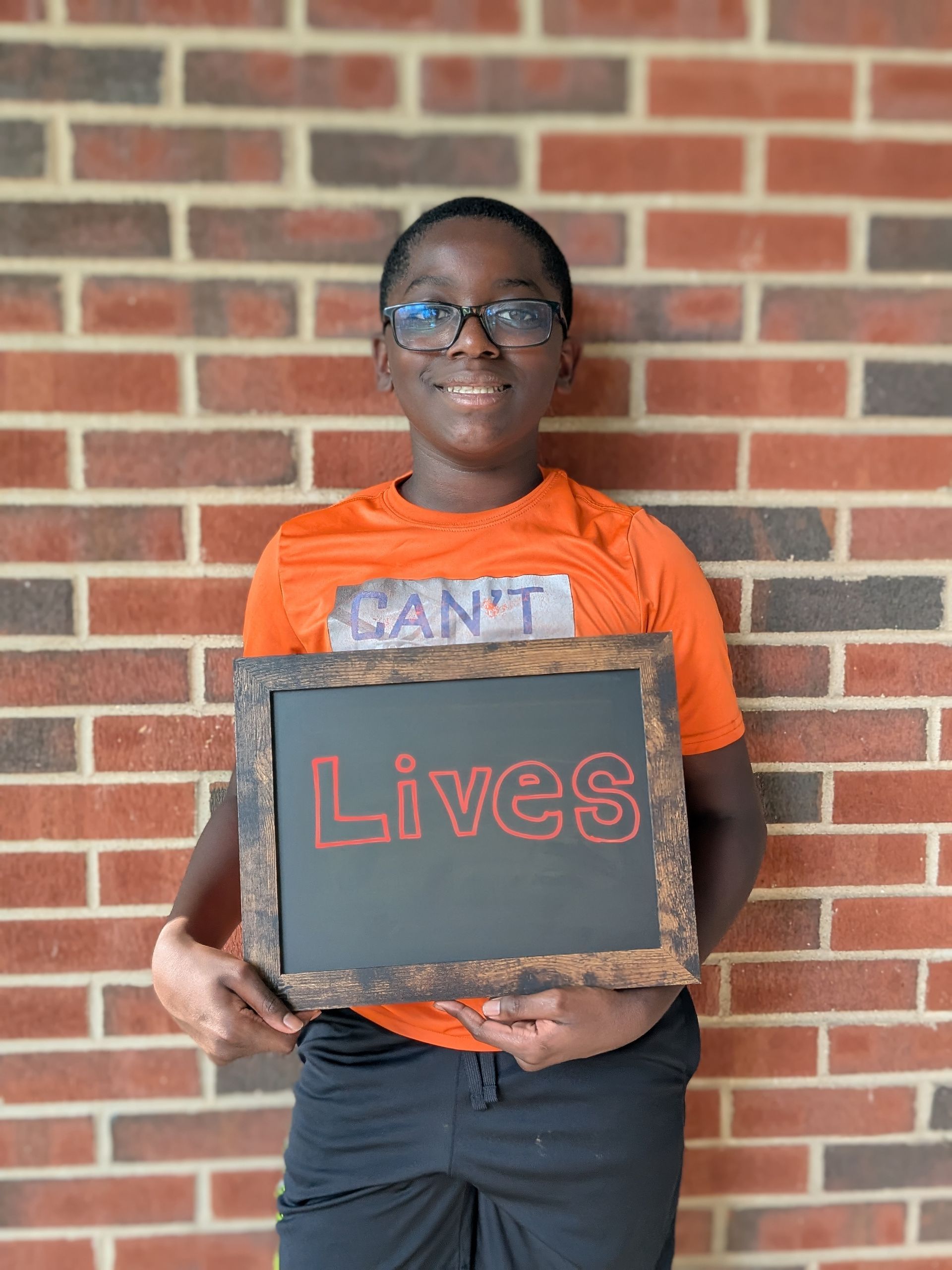 Young Black boy in orange shirt and glasses holds a chalkboard that reads 