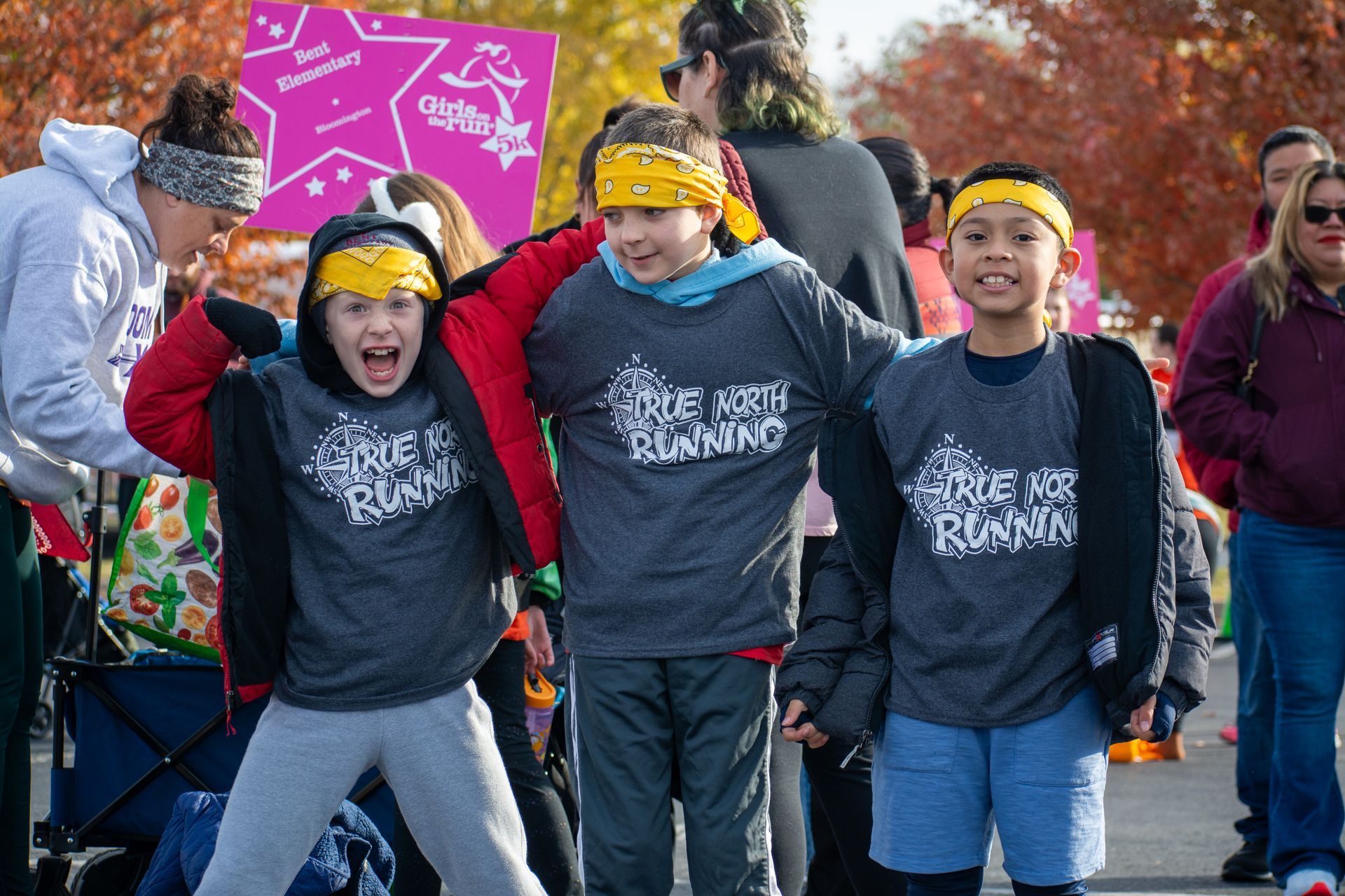 Three young boys in athletic clothes run on a sidewalk in front of a grassy area.