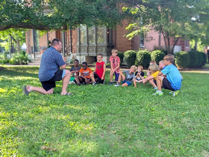 A group of children sit on a grassy lawn, listening to a kneeling adult. A brick building and trees are in the background.