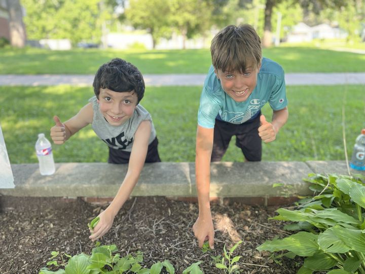 Two boys tending a garden, smiling and giving thumbs-up. They wear casual clothes and are outside on a sunny day.