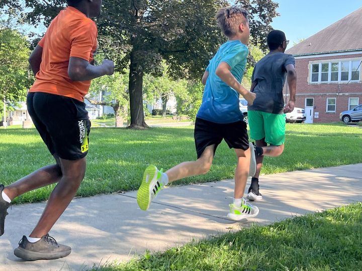 Three boys running on a sidewalk next to a grassy area. One wears orange, one blue, and the third green.