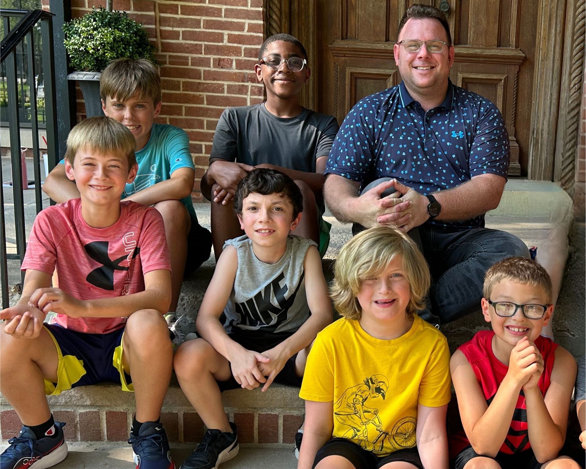 Group of boys and an adult man smiling and sitting on brick steps outside a building.