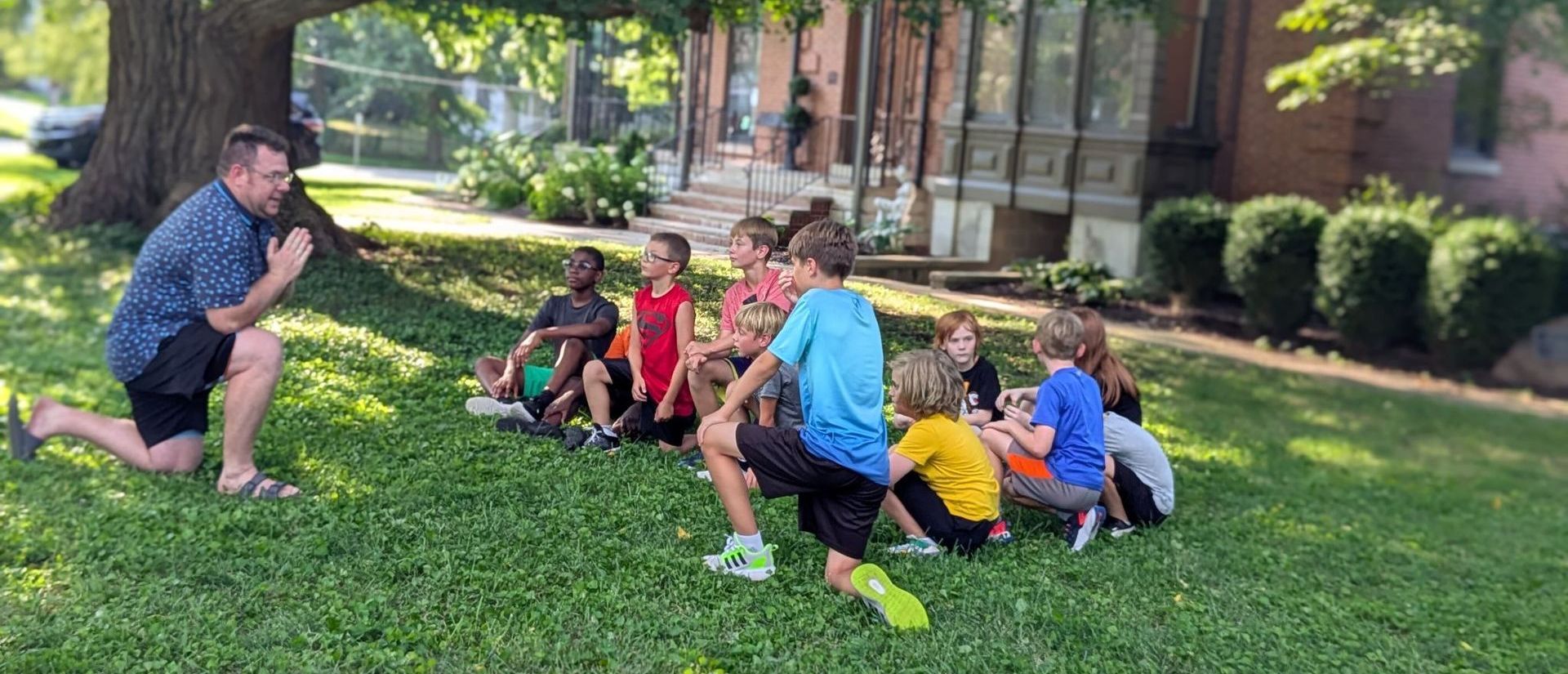 A man kneels in a grassy area, facing a circle of children. They are outdoors near a building and a large tree.
