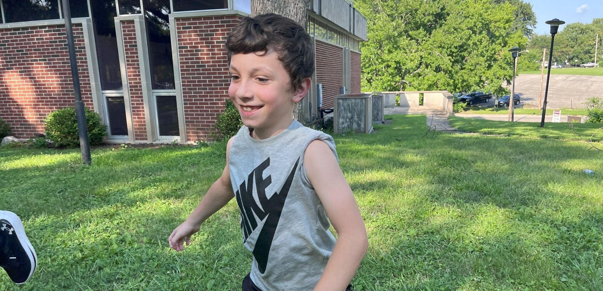 A young boy with curly hair and a gray Nike tank top smiles while running on a grassy lawn.