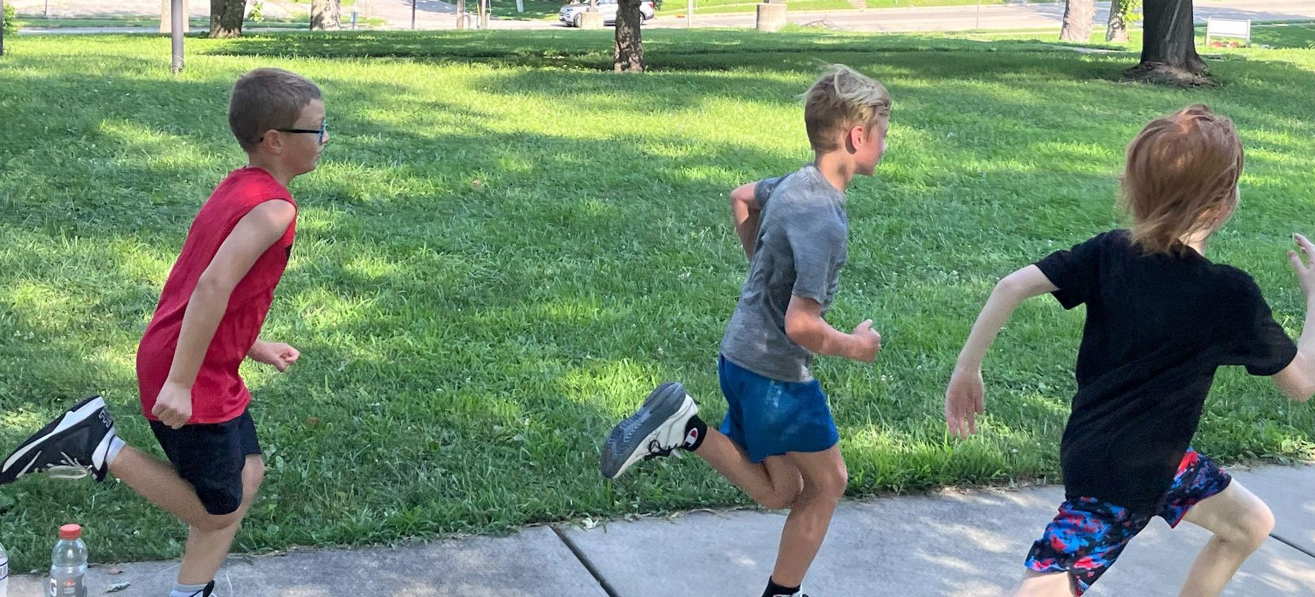 Three young boys in athletic clothes run on a sidewalk in front of a grassy area.