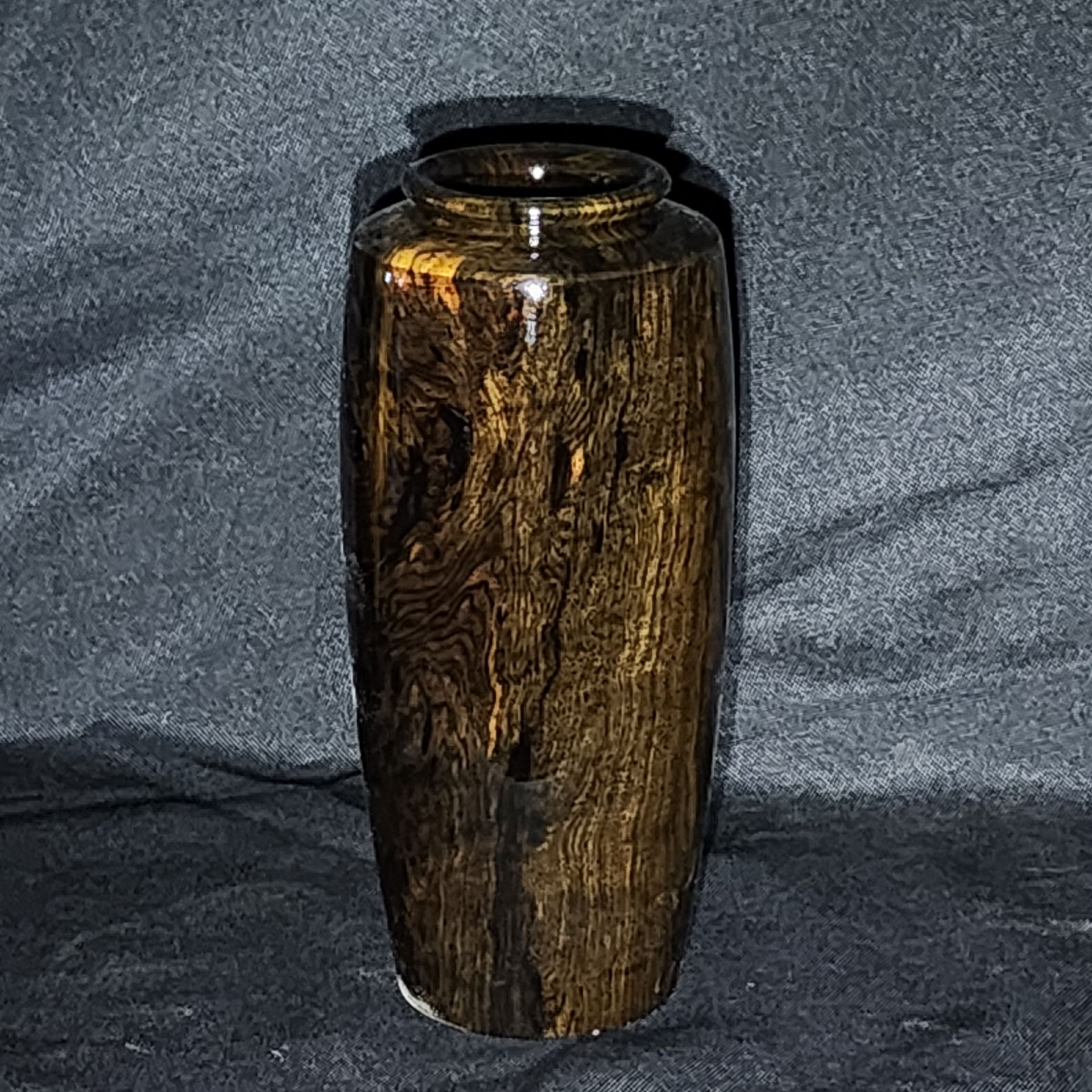 Tall, varnished wooden vase, standing upright on a dark, textured surface.