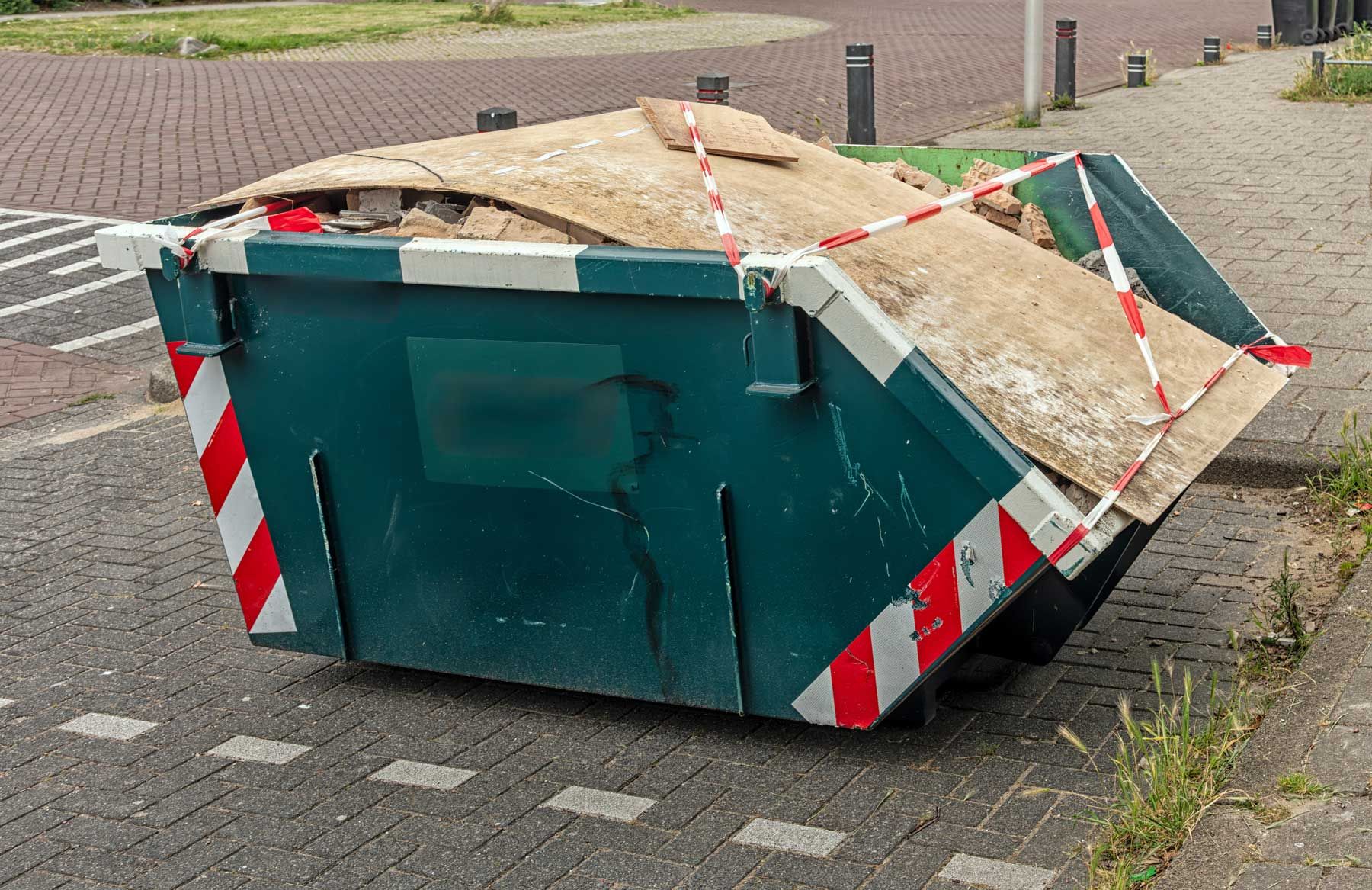 A full residential dumpster rental bin filled with construction debris on a brick driveway.