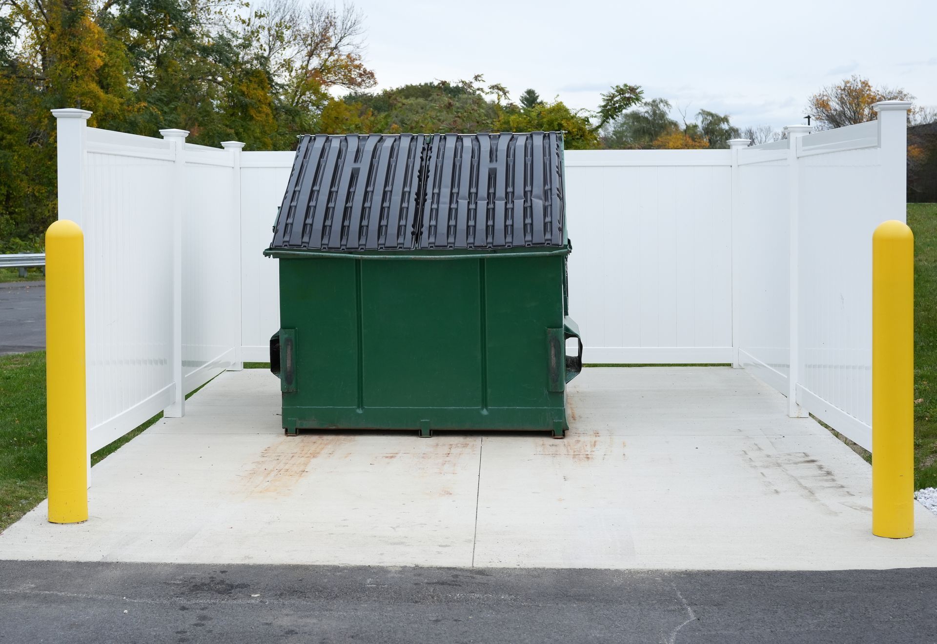 Green dumpster placed on a concrete pad, enclosed by white vinyl fencing with yellow bollards.