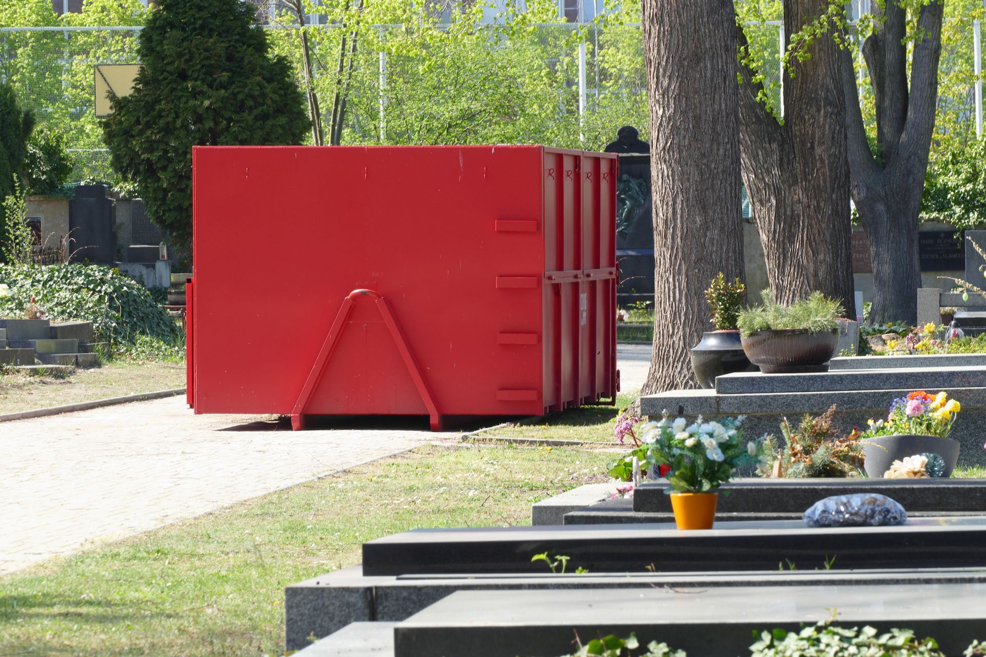 Red dumpster in cemetery with tombstones, flowers, and trees in background on paved path.