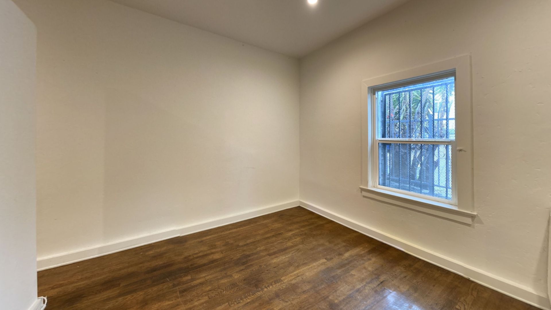 Empty room with hardwood floors, a window with security bars, and beige walls.