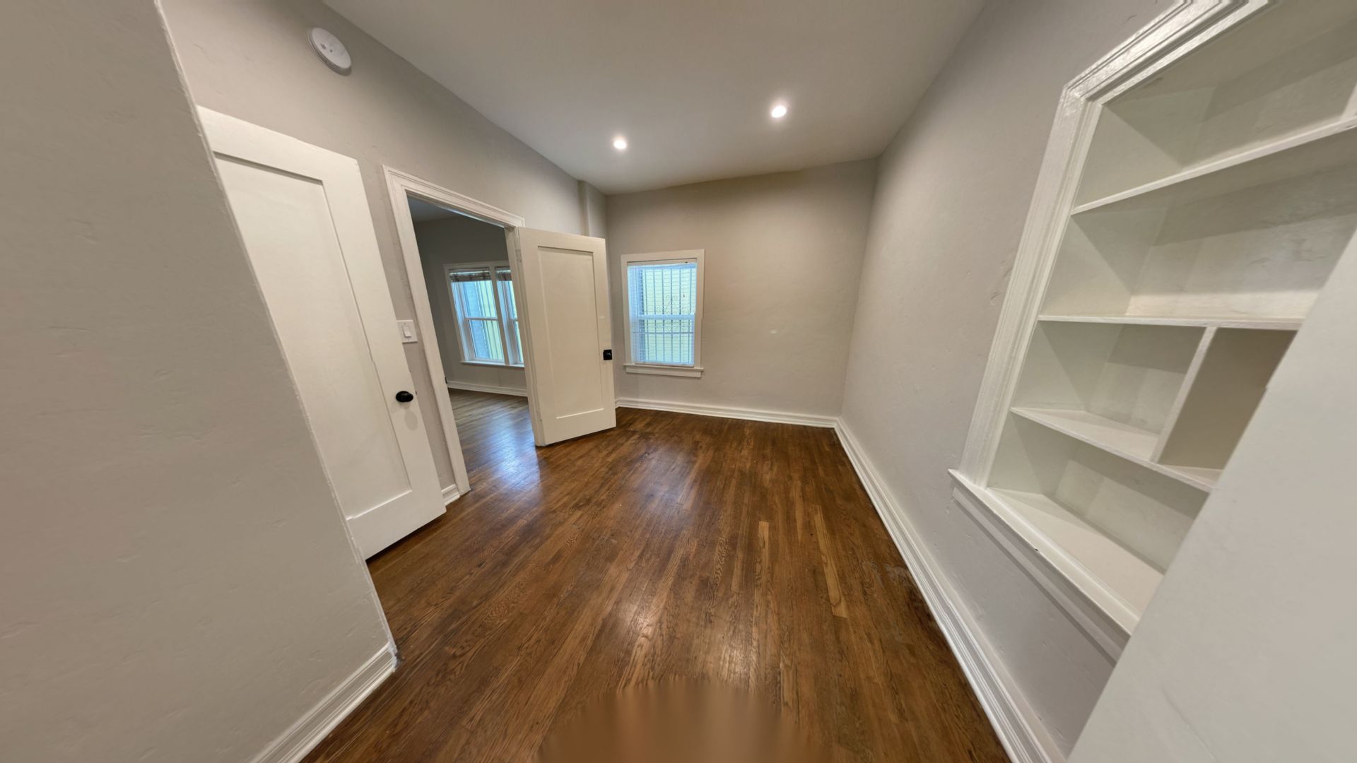Narrow room with hardwood floors, built-in shelves, window, and doorway to another room.