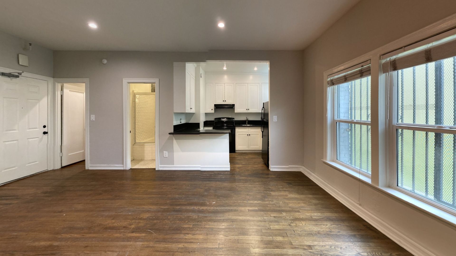 Living room with hardwood floors, white walls, and a view into the kitchen area. Two windows on the right side.