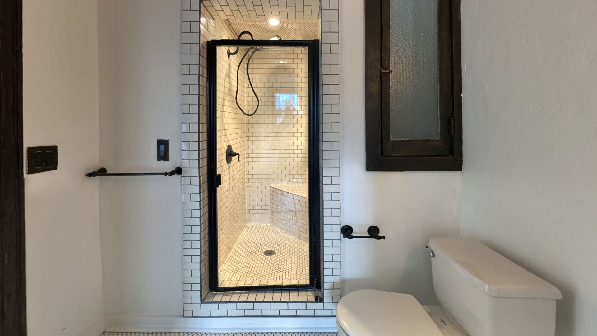 Small bathroom with white walls, shower with black frame, toilet, and window.