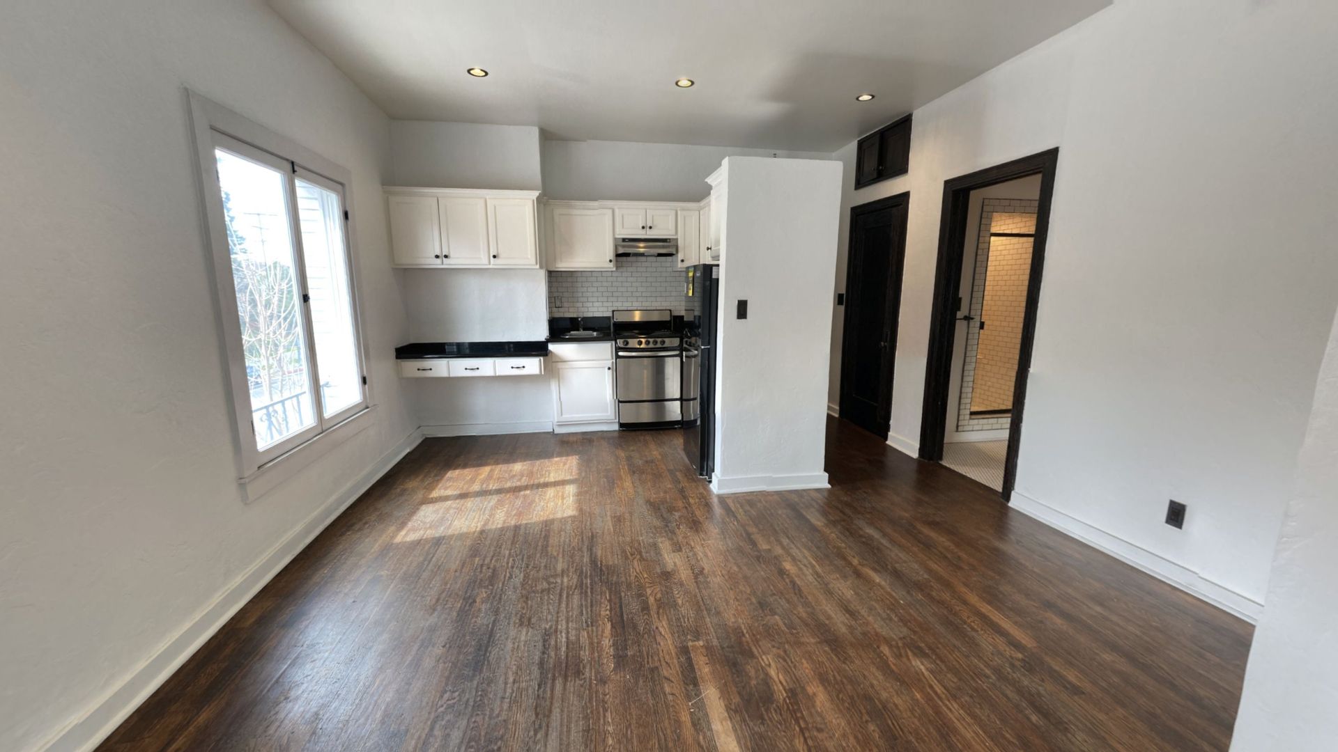 Small apartment interior with white walls, dark wood floors, kitchen, and doorway to a hallway.