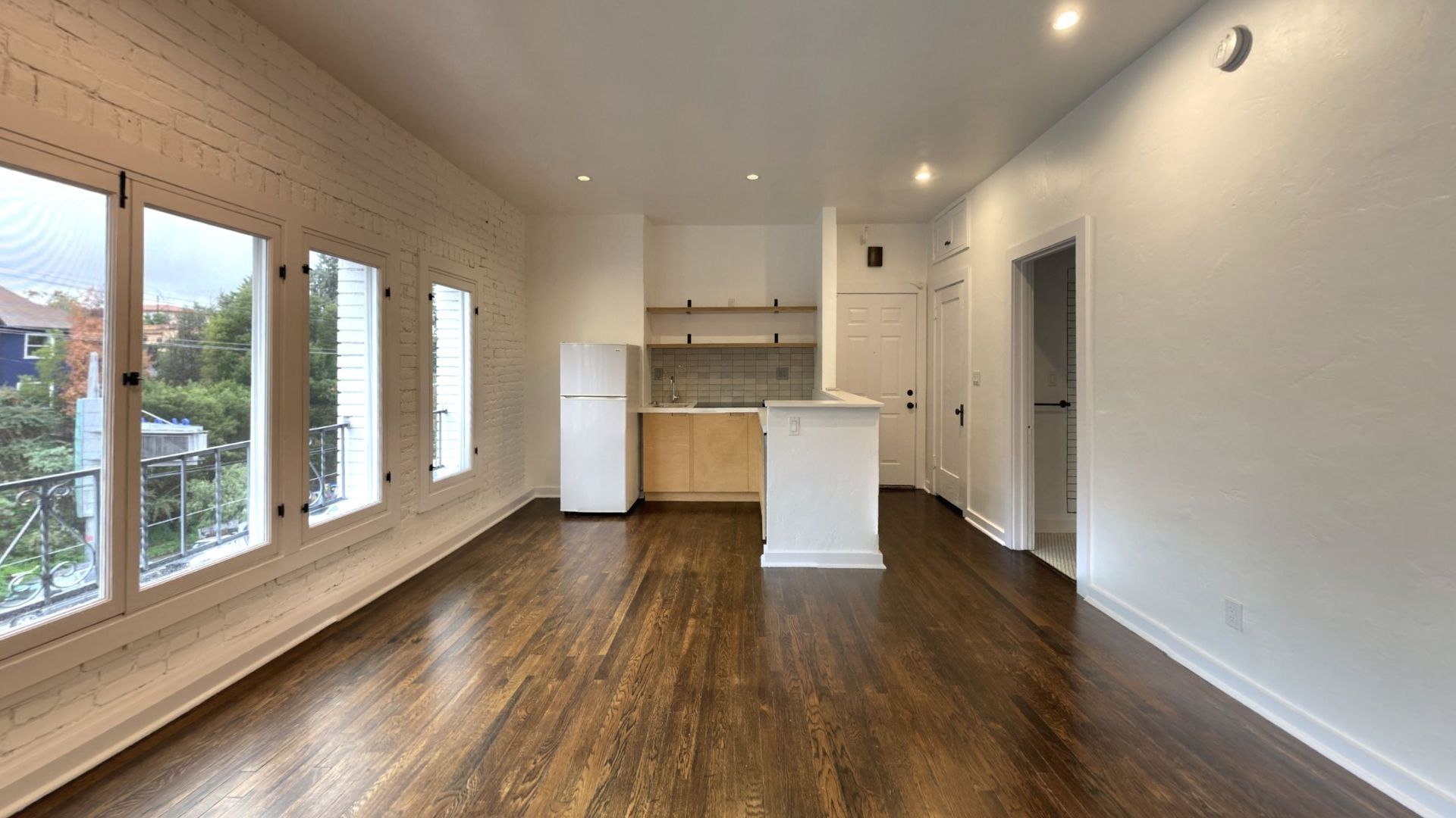 Empty apartment interior with dark wood floors, large windows, and a small kitchen.