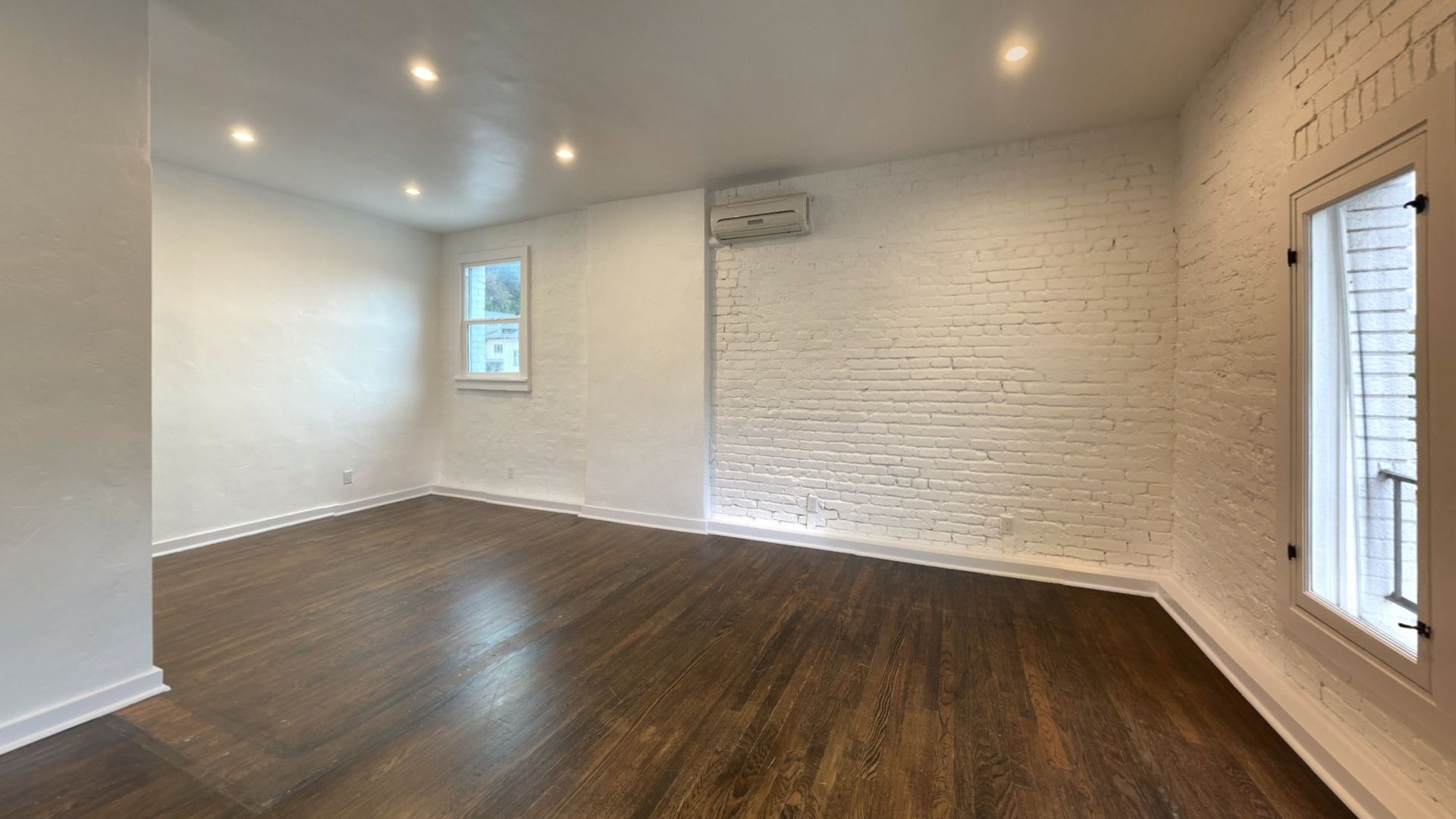 Empty room with hardwood floors, white walls, and exposed brick.