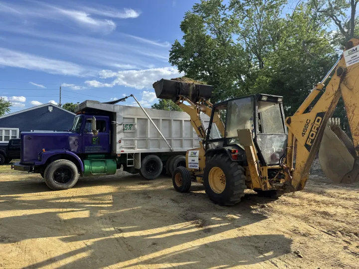 Yellow backhoe loader beside purple dump truck and white trailer in a gravel yard under a blue sky