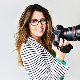 A woman wearing glasses and a striped jacket is holding a camera.