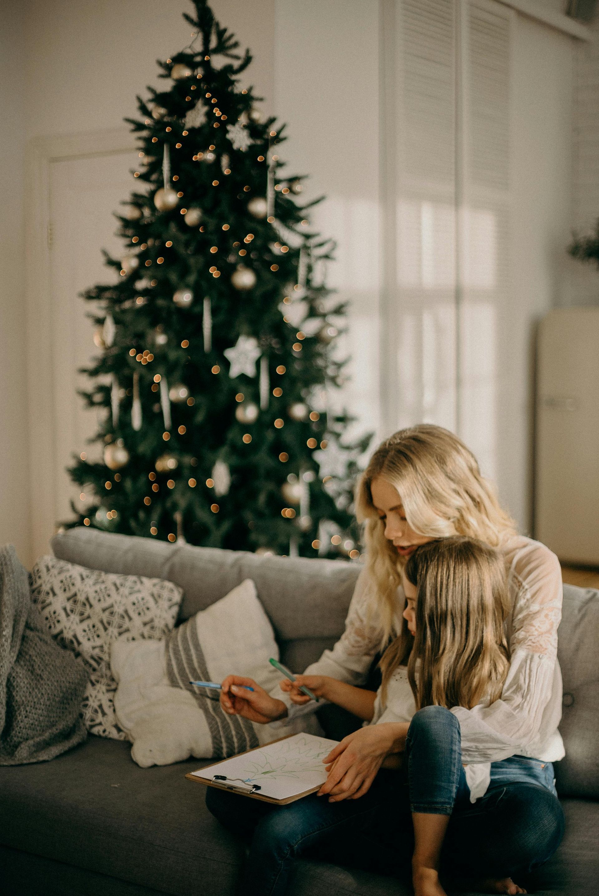 A mother and daughter are sitting on a couch in front of a christmas tree.