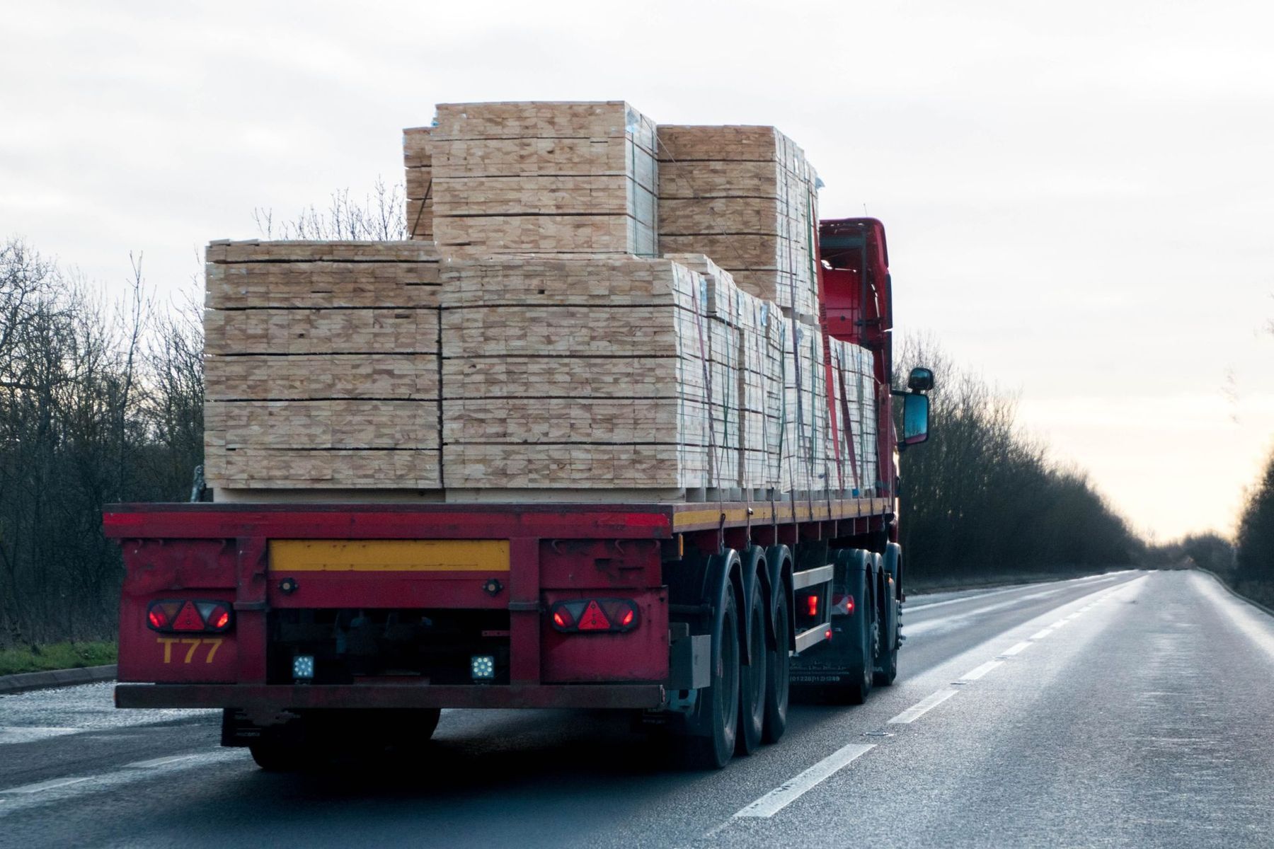 A red semi truck is driving down a highway with a stack of wood on the back.