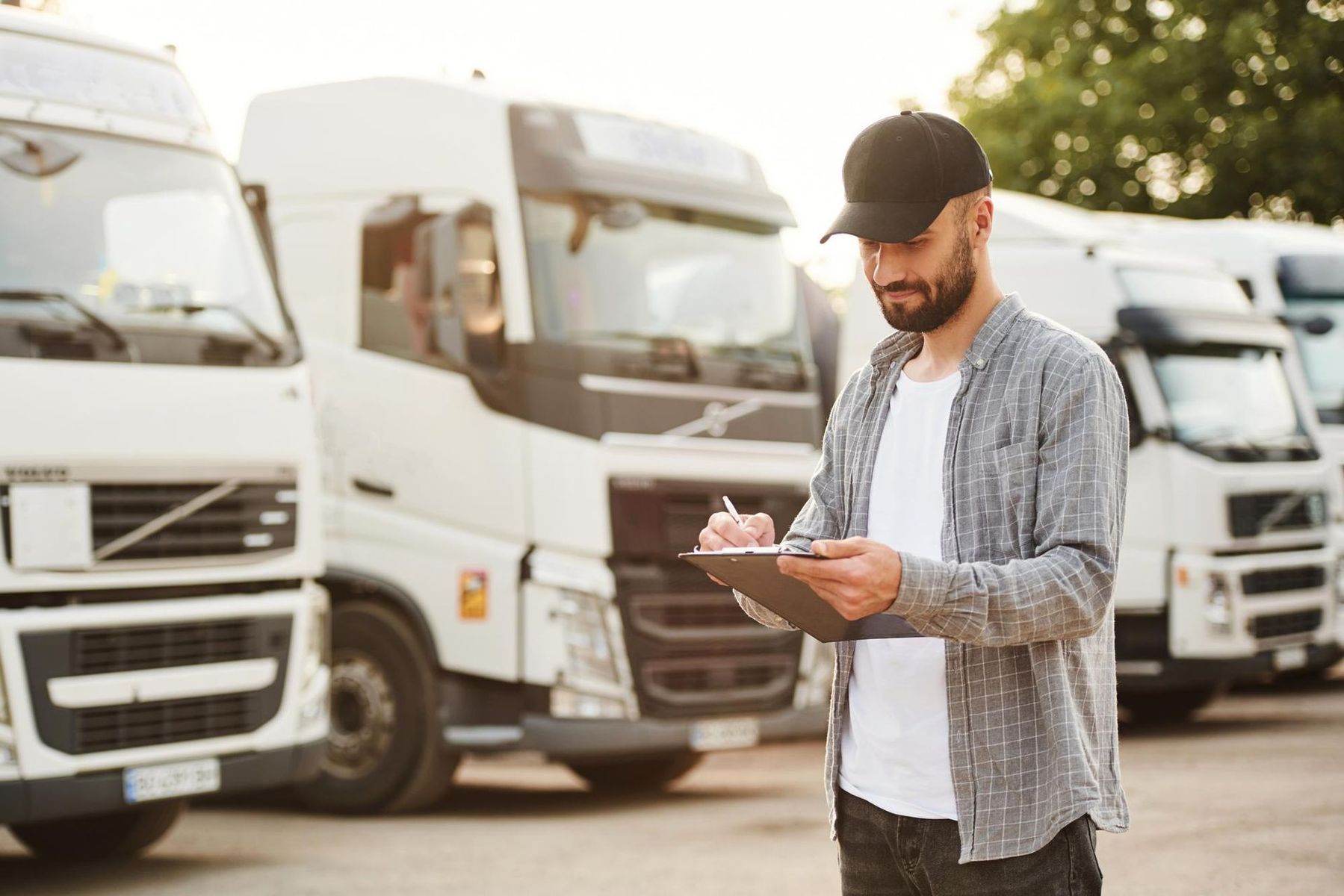 A man is standing in front of a row of semi trucks looking at a clipboard.