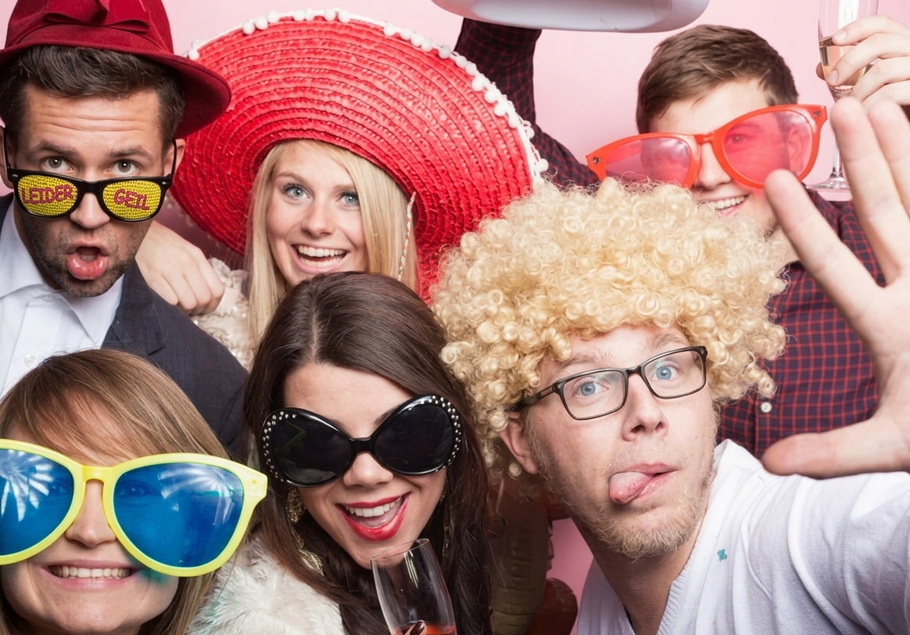 A group of people wearing colorful, silly props and oversized glasses pose joyfully for a photo against a pink background.