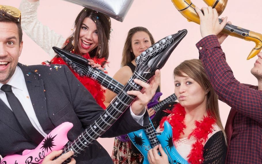 A group of people wearing party attire hold inflatable guitars and play, celebrating against a light pink background.