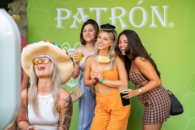 Four people posing for a photo in front of a green PATRÓN backdrop, smiling and holding drinks.