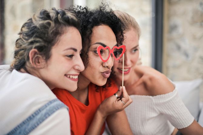Three people lean in closely together, smiling, while one holds heart-shaped novelty glasses in front of their face.